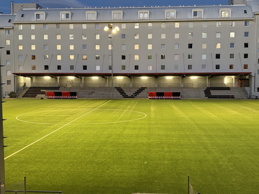 Der FavAC-Platz im Abendlicht mit beleuchteter Tribüne und den Häusern von Favoriten im Hintergrund